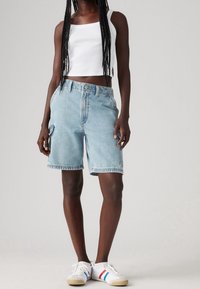 Mujer vestida con una camiseta sin mangas blanca, pantalones cortos de mezclilla azul claros hasta la rodilla con bolsillos tipo cargo, y zapatillas blancas con rayas rojas y azules.