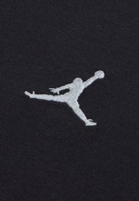 Svart stoff med en hvit, brodert Jumpman-logo som viser en figur i luften med ett ben utstrakt og armen hevet, med detaljert søm synlig.