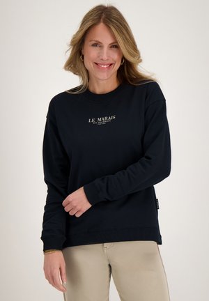 Lachende vrouw die een zwarte sweater met de tekst "Le Marais" en beige broek draagt, staand tegen een effen lichte achtergrond.