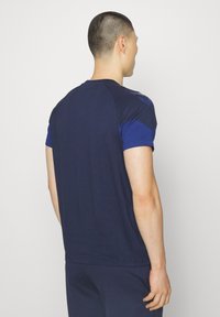 Mężczyzna z krótkimi włosami, zwrócony tyłem, ubrany w ciemnoniebieską koszulę z krótkim rękawem i pasujące ciemnoniebieskie spodnie na jednolitym jasnym tle.