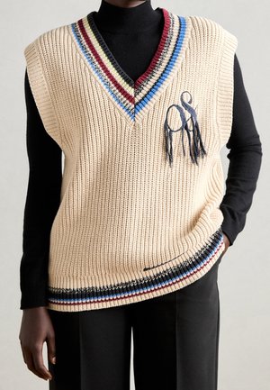 Person trägt eine cremefarbene, ärmellose Strickweste mit mehrfarbigen Streifen am Kragen und Saum über einem schwarzen Langarmshirt und schwarzen Hosen.