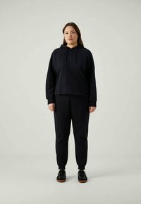 Frau, die steht, trägt einen schwarzen Kapuzenpulli, schwarze Jogginghosen und schwarze Sneakers mit braunen Sohlen vor einem schlichten hellen Hintergrund.
