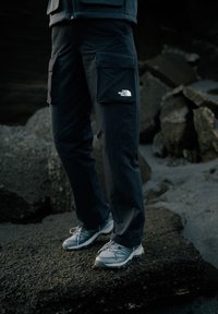 Schwarze Cargohosen mit großen Seitentaschen und einem weißen Logo am Oberschenkel, kombiniert mit grauen, schnürbaren Wanderschuhen mit strukturierten Sohlen.