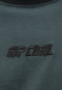 Κοντινή λήψη σκούρου πράσινου υφάσματος με μαύρο κεντημένο κείμενο "RIP CURL" με έντονα, στυλιζαρισμένα κεφαλαία γράμματα.