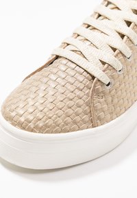 Beige pletené tenisky s texturovaným svrškem, vybavené bílými gumovými podrážkami a krémovými tkaničkami. Metalické detaily jemně září.