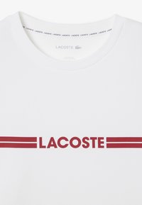 T-shirt bianco con collo a giro, testo rosso "LACOSTE" con due strisce orizzontali e una morbida texture del tessuto.