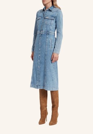 Femme portant une robe midi en denim bleu clair boutonnée, à manches longues et poches poitrine, associée à des bottes hautes en daim marron.
