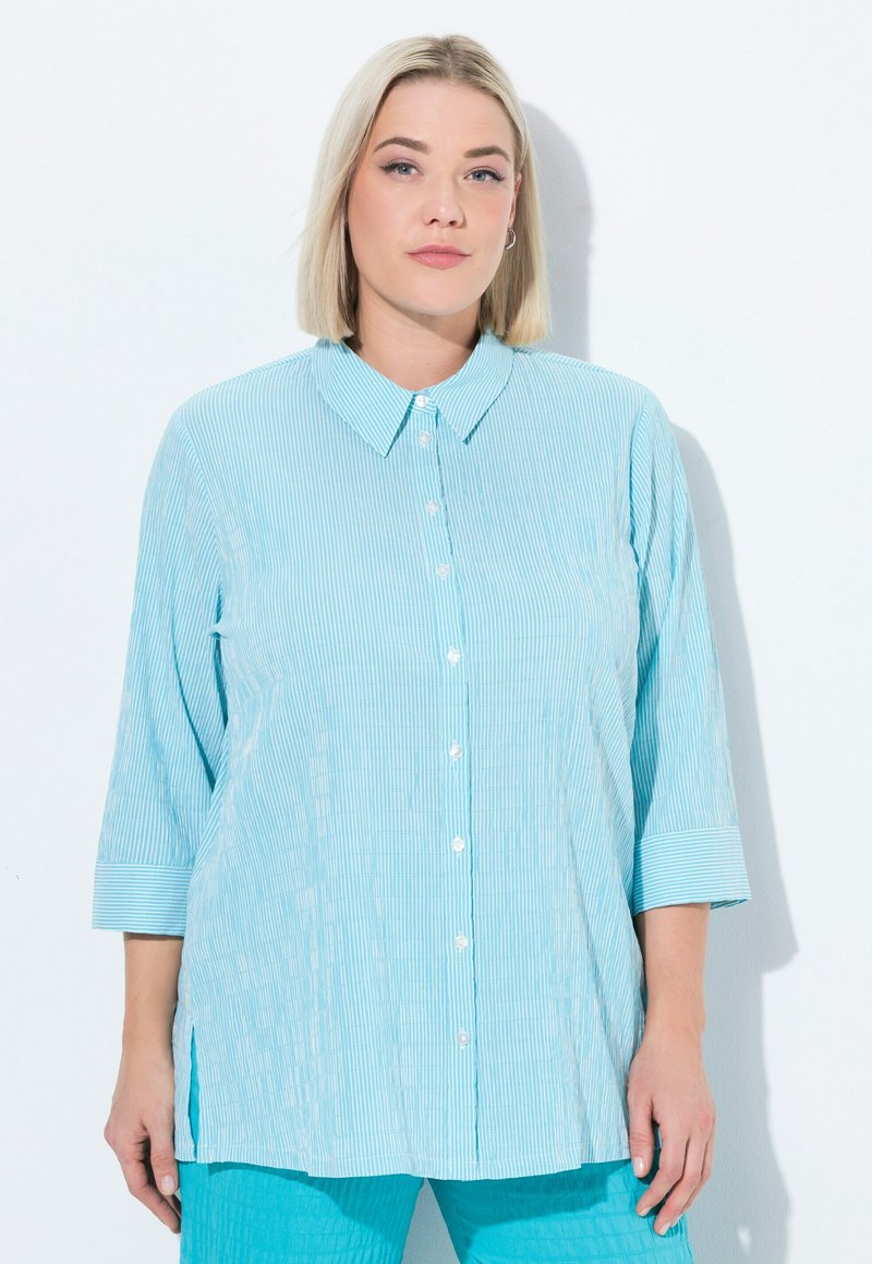 Camicia azzurro chiaro, con bottoni, a righe verticali, colletto e maniche a tre quarti. Presenta una vestibilità comoda e spacchi laterali per maggiore facilità.