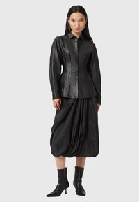 Giacca camicia in pelle nera con taglio sartoriale, chiusura a bottoni e maniche lunghe, abbinata a una gonna nera drappeggiata e stivaletti aderenti alla caviglia.