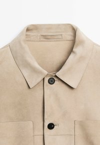 Beige suede jacket with a structured collar, two front pockets, and black buttons. Smooth texture with neat stitching details.