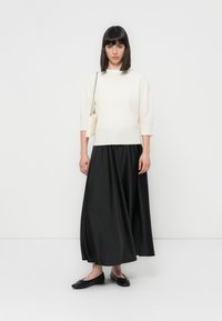 Femme avec des cheveux noirs lisses portant un pull blanc, une longue jupe noire, des ballerines noires et un sac blanc avec une bandoulière en chaîne.