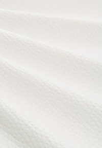 Witte stof met een gestructureerd patroon, voorzien van verheven stippen. Het oppervlak lijkt zacht en is in delicate plooien gedrapeerd.