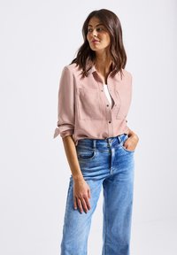 Žena oblečená v růžové košili na knoflíky s ohrnutými rukávy a modrých džínách s vysokým pasem, stojící na bílém pozadí.