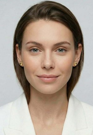 Ung kvinde med lys hud, glat brunt hår, grønne øjne, iført guldhjerteformede øreringe og en hvid blazer, smilende blidt.