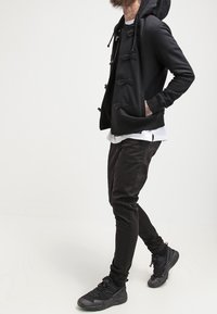 Sweat à capuche noir avec fermetures à cordon, superposé sur une chemise blanche. Associé à un pantalon noir ajusté et des baskets noires. Tissu lisse, design décontracté.