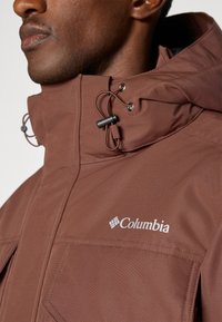 Rudas nepralaidus striukė su aukštu apykakle, reguliuojamais raišteliais ir ryškiu Columbia logotipu ant krūtinės. Tekstūruotas audinys.