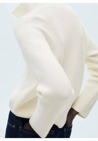 Cremfarbener Pullover mit gerippter Textur, hohem Kragen und weiten Ärmeln, kombiniert mit dunklen Jeans. Fokus auf Material- und Entwurfsdetails.