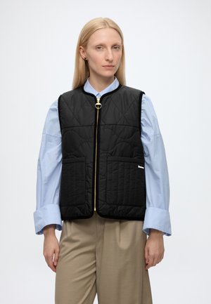 Femme portant un gilet matelassé noir avec des poches avant, par-dessus une chemise bleu clair et un pantalon beige, debout devant un fond blanc.