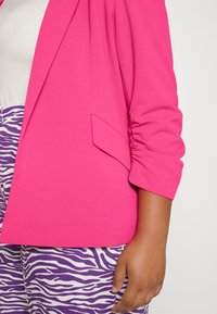 Blazer fucsia con maniche lunghe arricciate e una tasca singola, abbinato a pantaloni con stampa zebra viola. Texture liscia e vestibilità su misura.
