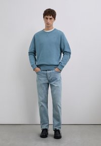 Joven de pie con las manos en los bolsillos, vistiendo una sudadera azul, jeans de un azul claro y zapatos negros, frente a una pared clara y simple.