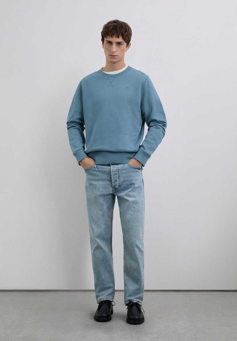 Joven de pie con las manos en los bolsillos, vistiendo una sudadera azul, jeans de un azul claro y zapatos negros, frente a una pared clara y simple.