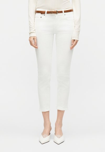 Γυναίκα που φοράει εφαρμοστά λευκά παντελόνια cropped, καφέ ζώνη, μπεζ τοπ με μακριά μανίκια και λευκά παπούτσια μυτερά με διακόσμηση δαντέλας.