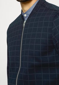 Uomo che indossa una giacca zip-up a scacchi blu scuro su una camicia azzurra con bottoni, che mostra il colletto e la parte superiore del petto.