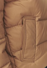 Bruine pufferjack met een geborduurde textuur, voorzien van een rechthoekige patch zak en zichtbare stiksels. Het materiaal lijkt glad en zacht.
