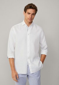 Camisa de lino blanca con cuello clásico y mangas arremangadas, que presenta un detalle a rayas azules contrastantes en el interior de los puños.