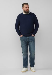 Navyblaues Langarmhemd mit strukturiertem Finish, kombiniert mit hellblauen Jeans und weißen Sneakers, vor einem neutralen Hintergrund stehend.