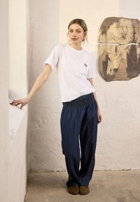 Femme portant un t-shirt blanc et un pantalon large bleu marine, debout à l'intérieur, s'appuyant légèrement sur un rebord de fenêtre blanc avec une œuvre d'art murale abstraite derrière elle.