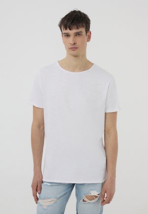 T-shirt bianca in lino a maniche corte con scollatura rotonda, caratterizzata da una vestibilità rilassata e una texture sottile, indossata con jeans azzurri chiari strappati.