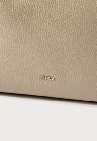 Beigenvärinen nahkamateriaali, jossa teksturoitu pinta; koristeltu kultaisella "Furla"-logolla. Pinnan hienovarainen rakenne.