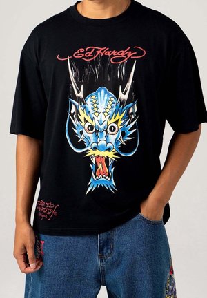 Czarny T-shirt z kolorową, groźną niebieską grafiką smoka, czerwonym tekstem "Ed Hardy" na górze, noszony z niebieskimi dżinsami.