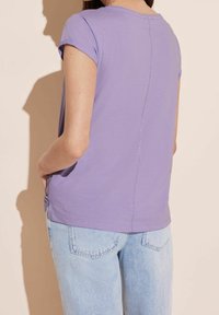 Nainen, joka käyttää lyhythihaista laventelinväristä paitaa ja vaaleansinisiä farkkuja, seisoo kädet taskuissa, kasvot poispäin yksinkertaista taustaa vasten.