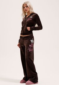 Frau mit langen blonden Haaren, die einen braunen Veloursjuicy Couture Trainingsanzug und pinke Hausschuhe trägt, steht vor einem schlichten hellen Hintergrund.