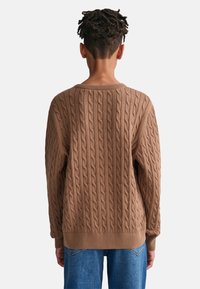 Suéter marrón de punto grueso con cuello redondo y puños acanalados, combinado con jeans de mezclilla azul, mostrando un patrón texturizado en la tela.