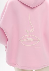 Rosa Kapuzenpullover mit einem gestickten Linienbild von Lippen und Nase in Creme, mit lockerem Schnitt und weicher Textur.