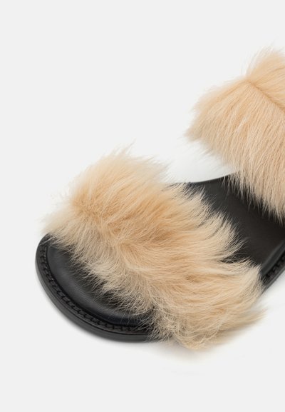 Black slide sandal featuring two thick, fluffy beige faux fur straps, a smooth leather footbed, and a contrasting stitched edge detail.