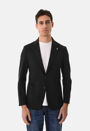 Uomo che indossa un blazer nero con due bottoni, t-shirt bianca e jeans blu, in piedi davanti a uno sfondo chiaro e semplice.