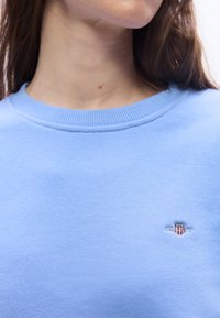 Άτομο που φοράει γαλάζιο φούτερ με στρογγυλή λαιμόκοψη και μικρό κεντημένο έμβλη μα σε κόκκινο, άσπρο και μπλε στο στήθος.