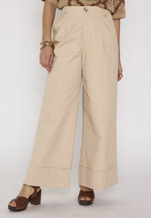 Pantaloni beige a gamba larga con bottone e pieghe, abbinati a sandali marroni con tacco alto e braccialetti dorati al polso sinistro.