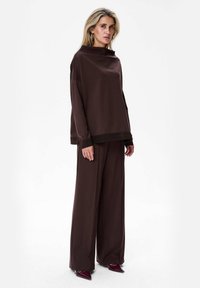 Brązowy, oversize top z wysokim dekoltem i prążkowanym wykończeniem, w połączeniu z szerokimi brązowymi spodniami. Lśniące burgundowe, spiczaste buty. Gładka faktura.