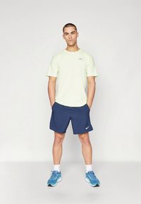 Svetložltá košeľa s krátkym rukávom a malým logom, kombinovaná s námornícky modrými šortkami a modrou športovou obuvou, zobrazená na neutrálnom pozadí.