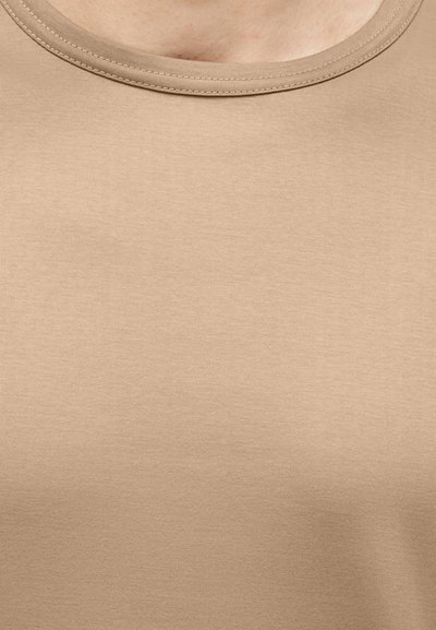 T-shirt beige à col rond porté par une personne, montrant le haut de la poitrine et la zone du cou.