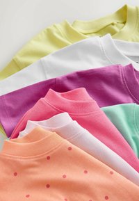 T-shirts à col rond superposés en jaune pastel, blanc, violet, rose, vert menthe et pêche avec des pois roses, disposés en un agencement ordonné et superposé.