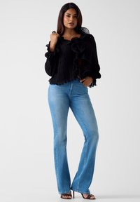 Czarna plisowana bluzka z falbankami na kołnierzu i rękawach, zestawiona z jasnoniebieskimi dzwonowymi dżinsami. Modelka stoi na jednolitym tle.