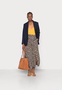 Blazer azul-marinho sobre blusa mostarda, saia com padrão floral e fenda lateral, mala bege e sandálias de salto bloco em tom bege claro.