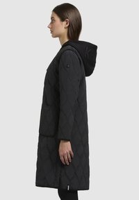 Cappotto trapuntato nero con un design lungo e dritto. Presenta un cappuccio, tasche laterali e un motivo testurizzato. Il tessuto sembra liscio e caldo.