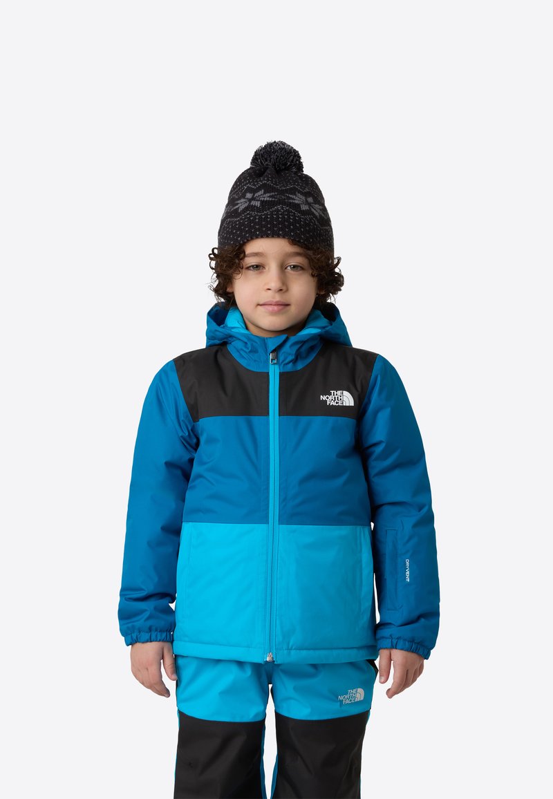 Modrá a čierna zateplená bunda s kapucňou, s veľkými farebnými blokmi. Dieťa má na sebe zodpovedajúci snehovo modrý outfit a čiernu pletenú čiapku.